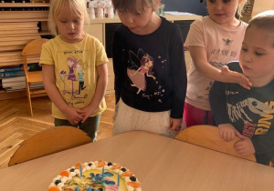 6 urodziny Zosi. Na stoliku jest tort z zapalonymi świeczkami. Zosia dmucha na świeczki. Obok niej stoi trójka dzieci.